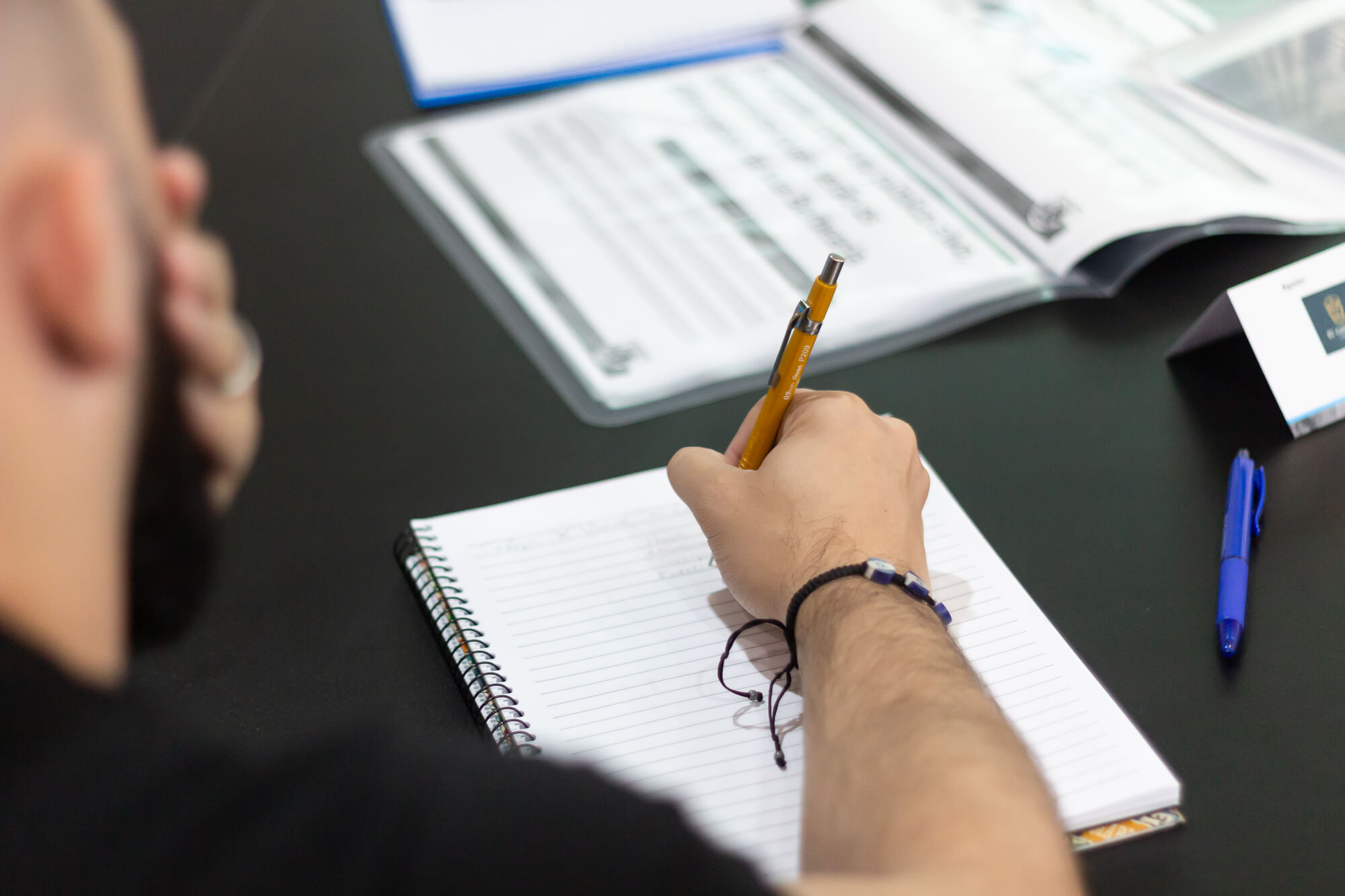 Mão escrevendo com caneta em um caderno de anotações durante uma reunião de planejamento.