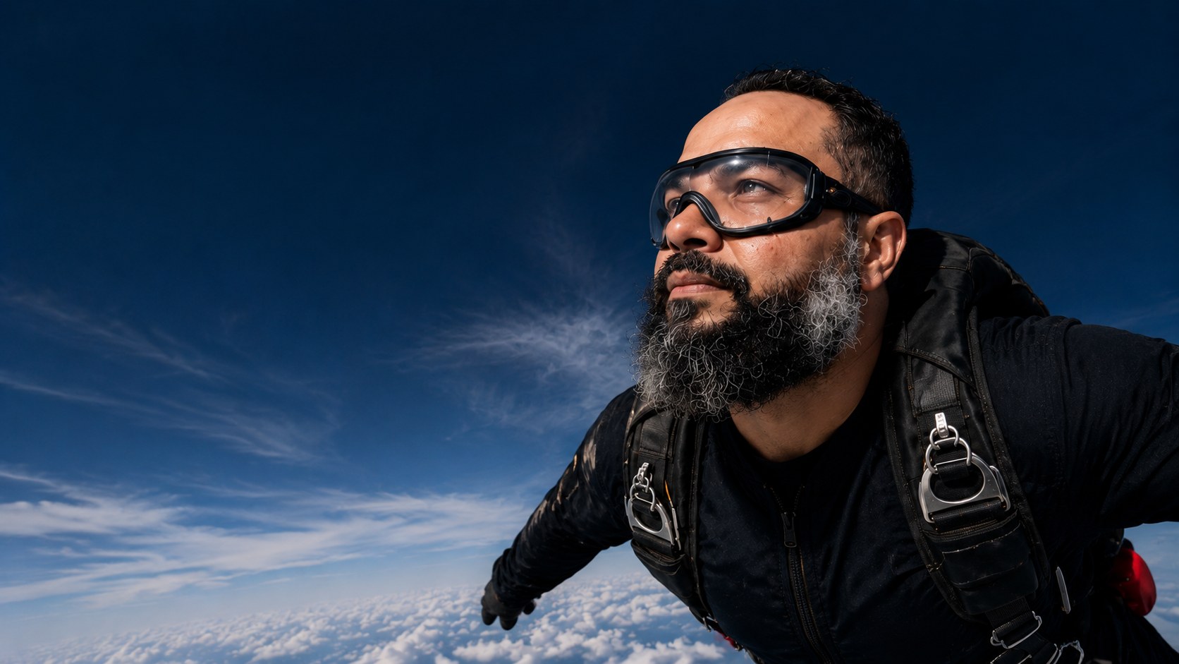 Paraquedista em queda livre olhando para o horizonte, céu azul profundo e nuvens abaixo.