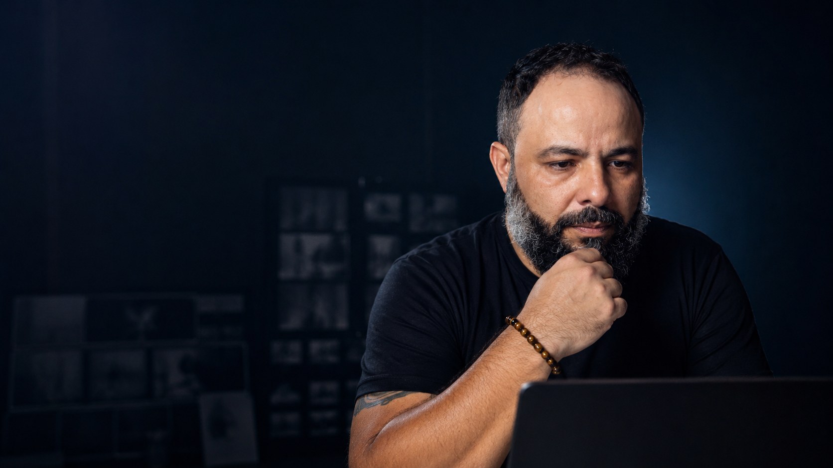 Dennis Phillipps pensativo diante do notebook, com fotos de trabalho na parede ao fundo, em luz baixa.